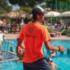 Persona con maglietta arancione in piscina, diverse persone nuotano.